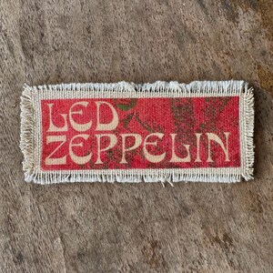 May include: A rectangular fabric patch with frayed edges featuring the text LED ZEPPELIN in a cream-colored serif font. The background of the patch is a textured red pattern with dark green accents visible behind the lettering.