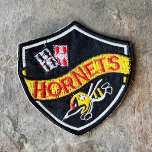 May include: A shield-shaped embroidered patch with a black background and a white border. It features a yellow banner across the center with the red text HORNETS, a white and red playing card graphic, and a yellow and white hornet illustration.
