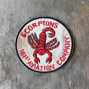 May include: A circular embroidered patch with a white background and a black border. The center features a red scorpion graphic. The text SCORPIONS COMPANY is arched across the top, and 161ST AVIATION COMPANY is arched along the bottom in red.