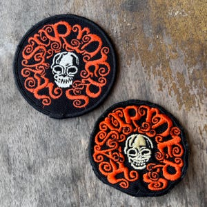 May include: Two circular embroidered patches featuring a black background with a central white skull surrounded by orange swirling text that reads "SURRENDER". The patches are identical in design, with one being slightly larger than the other.