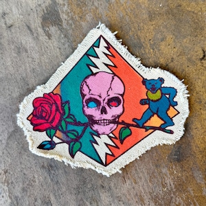 May include: A diamond-shaped fabric patch with frayed edges featuring a pink skull holding a red rose in its teeth. A blue bear stands to the right of the skull. The background consists of diagonal teal and orange stripes with white lightning bolts.