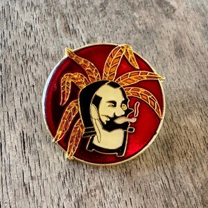 May include: A round, red and gold enamel pin featuring a stylized face with a black beard and hair, and a tongue sticking out. The pin has gold accents and a gold border.