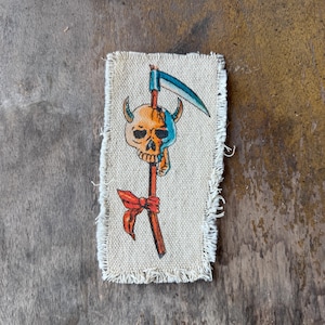 May include: A rectangular beige fabric patch with frayed edges featuring a hand-drawn illustration of a horned skull split between orange and blue colors. A scythe blade extends from the top, and a red ribbon is tied around the handle below.