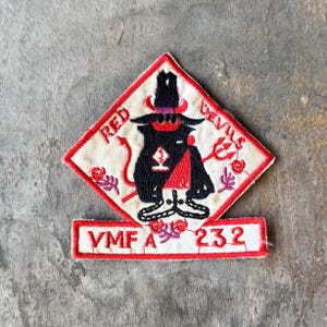 May include: A diamond-shaped embroidered patch with a white background and red border. It features a black devil figure wearing a hat, with the text RED DEVILS. Below, a rectangular tab displays the red embroidered text VMF A 232.