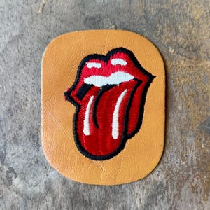 May include: A tan leather patch featuring an embroidered design of a red mouth with white teeth and a tongue. The design is outlined in thick black thread, creating a bold graphic contrast against the smooth, light brown leather background.