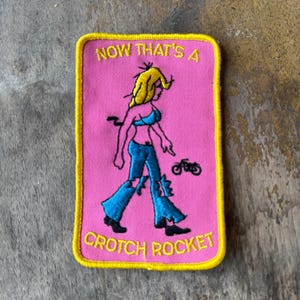 May include: A rectangular embroidered patch with a pink background and a yellow border. It features a yellow-haired figure in a blue top and blue jeans, a small black motorcycle icon, and the yellow text "NOW THAT'S A CROTCH ROCKET".