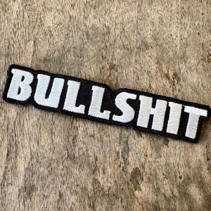 May include: A black embroidered patch with the word "BULLSHIT" in white, angled across the patch. The patch has a black border and is set against a weathered wooden surface.