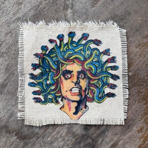 May include: A square fabric patch with a frayed edge featuring a colorful graphic print of a Medusa figure. The central character has a pale face with wide eyes and an open mouth, surrounded by a dense mass of blue and yellow snakes for hair.