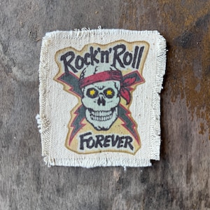 May include: A square fabric patch with frayed edges featuring a graphic of a skull wearing a red bandana. Behind the skull are two crossed dark lightning bolts. The text Rock'n'Roll is printed above the skull and FOREVER is printed below.