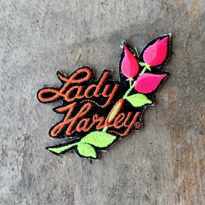 May include: An embroidered patch featuring the text Lady Harley in orange cursive script on a black background. The design includes two bright pink flower buds and light green leaves extending from the right side of the lettering.