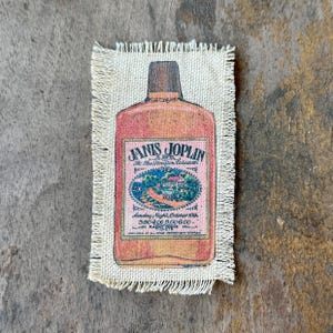 May include: A rectangular piece of off-white fabric with frayed edges featuring a printed graphic of a liquor bottle. The label reads Janis Joplin at the Houston Coliseum, Sunday Night, October 19th, 3.50, 4.00, 5.00, 6.00, Radio KNUZ tickets.