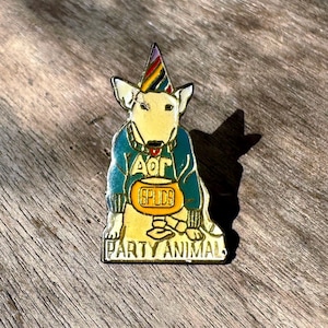 May include: A colorful enamel pin featuring a cartoon Bull Terrier wearing a party hat and a blue sweater. The pin has the text "PARTY ANIMAL" and a "SPUDS" design. The pin is yellow, blue, green, and red.