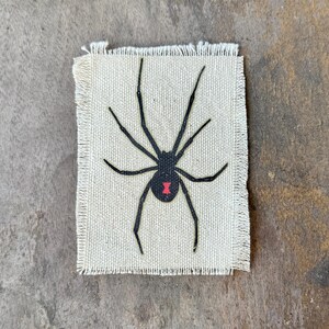 May include: A rectangular off-white fabric patch with frayed edges featuring a black spider illustration centered on the surface. The spider has eight long, thin legs and a small, distinct red hourglass-shaped marking on its rounded abdomen.