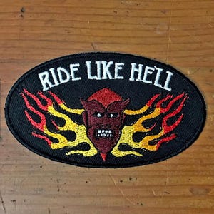 May include: Black oval patch with the text "RIDE LIKE HELL" in white. The patch features a red devil face with flames in yellow and orange. The patch is likely for a motorcycle jacket or vest.