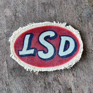 May include: An oval-shaped fabric patch with a frayed, off-white border. The center features a deep red background with the letters LSD printed in a large, bold, light-colored font with dark blue outlines, creating a distinct visual contrast.