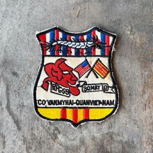 May include: A shield-shaped embroidered patch featuring a red devil figure, two crossed flags, and the text RPG59, 30MAT10, and COVANMYHAI-QUANVIETNAM. The design includes red, white, blue, and yellow stripes with a black border on a white background.
