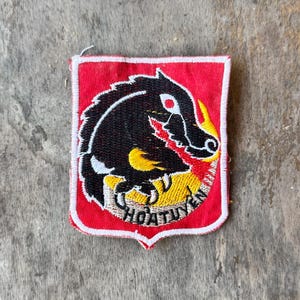 May include: A rectangular embroidered patch with a red background and a white border. The design features a black stylized animal head with yellow accents, a red eye, and the text HOATUYEN stitched in black at the bottom of the emblem.
