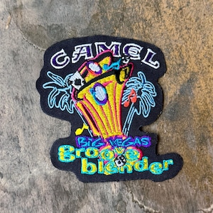 May include: A black embroidered patch featuring the text CAMEL at the top and BIG VEGAS groove blender at the bottom. The design includes a yellow cup with pink outlines, blue palm trees, musical notes, and a small die graphic in the center.