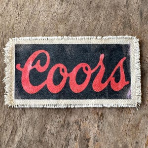 May include: A rectangular fabric patch featuring a black background with the word Coors printed in a red cursive script font. The patch has a light beige border and frayed edges on all four sides, creating a textured and rustic appearance.