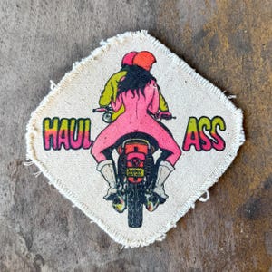 May include: A square-shaped fabric patch with frayed edges featuring a graphic illustration of a person riding a motorcycle. The person has dark hair and wears a pink bodysuit. The text HAUL ASS is printed in a colorful gradient font.