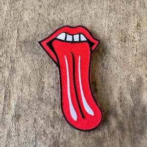 May include: A fabric patch featuring an embroidered design of a pair of red lips with an extended tongue. The design includes white teeth at the top and two vertical white lines running down the center of the tongue, all outlined in black thread.