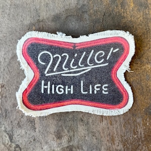 May include: A rectangular fabric patch with frayed edges featuring a black background and a red border. The text Miller is written in a white cursive script, and the text HIGH LIFE is printed below in a white sans-serif font.
