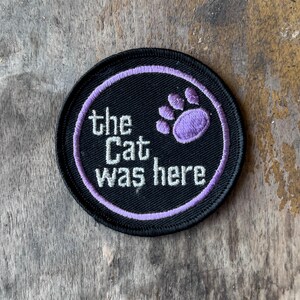 May include: A circular embroidered patch with a black background and a lavender border. The patch features white text that reads "the Cat was here" and a lavender paw print graphic positioned to the right of the text, all rendered in thread.