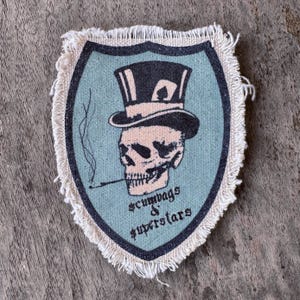 May include: A shield-shaped fabric patch with a frayed edge featuring a light blue background and a dark border. The design shows a skull wearing a black top hat with a playing card detail, smoking a cigarette, and the text scumbags & superstars.