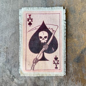May include: A rectangular fabric patch with a frayed edge featuring a printed design of an ace of spades playing card. The graphic shows a skull holding a scythe centered within a large black spade symbol, with small crossbones and spade icons.