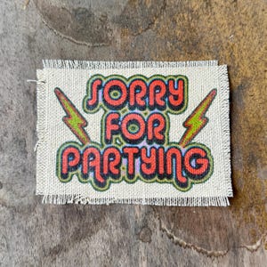 May include: A rectangular fabric patch with frayed edges featuring the text SORRY FOR PARTYING in bold red letters with black and blue outlines. Two green lightning bolt graphics are positioned on the left and right sides of the text.