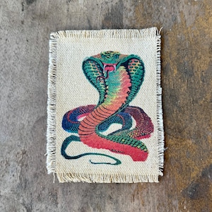 May include: A rectangular beige fabric patch with frayed edges featuring a colorful illustration of a cobra snake. The snake is depicted with a flared hood, a red and orange underbelly, and green and blue scales, with its mouth open showing fangs.