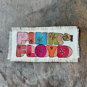 May include: A rectangular fabric patch with frayed edges featuring the text PINK FLOYD in large, multi-colored, bubble-style letters. Each letter contains a small flower graphic, and a hand icon points toward the right side of the text.