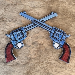 May include: Embroidered patch featuring two crossed revolvers. The pistols have blue bodies, black detailing, and red grips. The patch is designed to be sewn onto clothing or accessories.