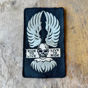 May include: A rectangular black embroidered patch featuring a central white skull with wings extending upward and downward. The text RIDE TO LIVE and LIVE TO RIDE is stitched in white on either side of the skull, all enclosed by a black border.