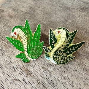 May include: Two enamel pins featuring a cobra design. The pins have a green and gold color scheme, with a cobra head and body, and green leaf-like shapes around the snake. The cobra's eyes are red, and the background is a wooden surface.
