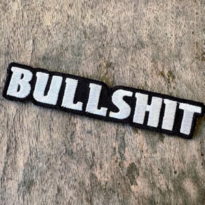 May include: A rectangular embroidered patch featuring the word BULLSHIT in bold, white, block-style capital letters set against a solid black background. The embroidery has a visible textured thread pattern throughout the lettering and border.