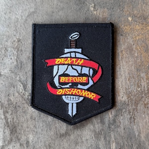 May include: A black shield-shaped embroidered patch featuring a white skull and a vertical sword. A red ribbon banner wraps around the skull and sword with the yellow embroidered text DEATH BEFORE DISHONOR displayed across the banner segments.