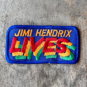 May include: A rectangular embroidered patch with a blue background. The text JIMI HENDRIX is stitched in yellow at the top. Below, the word LIVES is embroidered in large, bold letters featuring a gradient of red, yellow, and green thread colors.