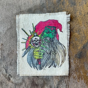 May include: A rectangular fabric patch with frayed edges featuring a printed illustration of a green-skinned figure with a long beard wearing a red hood. The figure holds a glowing orb containing a black skull graphic against a light beige background.