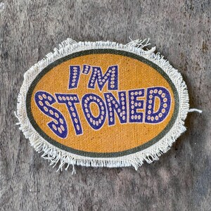 May include: An oval-shaped fabric patch with a mustard yellow background and a dark green border. The text I'M STONED is printed in dark purple with a dotted pattern. The patch features a frayed, off-white fringe edge around the entire perimeter.