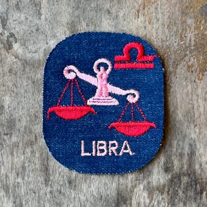 May include: A dark blue embroidered patch featuring a light pink scale of justice icon with bright red hanging pans and a red horizontal bar symbol above. The word LIBRA is stitched in light pink capital letters at the bottom of the patch.