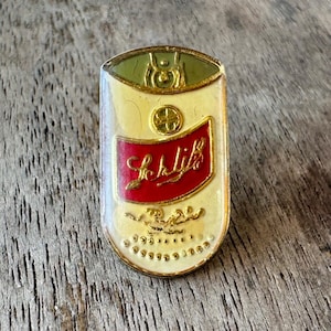 May include: Vintage Schlitz beer pin. The pin is a rectangular shape with rounded edges. The top is green, the middle is cream-colored with a red banner that reads "Schlitz" in gold script. The bottom has a cream-colored border with gold dots.