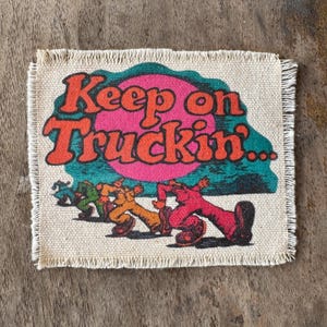 May include: A rectangular fabric patch with frayed edges featuring the text Keep on Truckin in orange letters. The design includes a large pink circle, a teal background shape, and a series of figures walking in red, orange, and green colors.