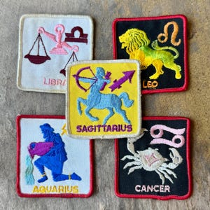 May include: Five square embroidered patches featuring zodiac signs. Libra in pink on white, Leo in yellow on black, Sagittarius in blue on yellow, Aquarius in blue on white, and Cancer in white on black. Each patch includes the sign name in text.