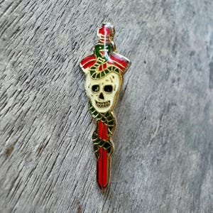 May include: A gold-colored enamel pin featuring a skull with a snake wrapped around a red dagger. The skull has a cream-colored face with black eye sockets and a grinning mouth. The snake is green and red, and the dagger is red.