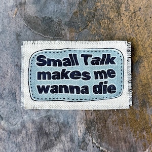 May include: A rectangular fabric patch with frayed edges featuring a light blue center panel with a dark blue stitched border. The text "Small Talk makes me wanna die" is printed in bold, dark blue, rounded sans-serif lettering across the center.