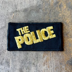 May include: A rectangular black embroidered patch featuring the text THE POLICE stitched in a bold, golden yellow thread. The lettering is arranged in two lines with a slight upward slant, and the entire patch has a finished black border.