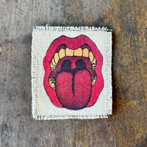 May include: A square fabric patch with frayed edges featuring a graphic illustration of an open mouth with bright red lips, yellow teeth, and a large, prominent red tongue centered on a light beige, textured, woven canvas background.