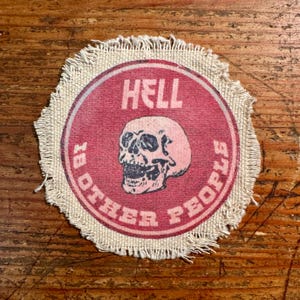 May include: A round patch with a red center featuring a skull graphic and the text "HELL IS OTHER PEOPLE" in white. The patch has a frayed edge and is set against a wooden background.