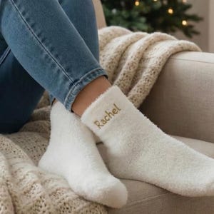 Peut inclure: Scène chaleureuse avec une personne sur un canapé beige, portant un jean bleu et des chaussettes blanches duveteuses brodées du nom "Rachel". Une couverture crème ajoute de la chaleur. Un sapin de Noël et une cheminée créent une ambiance festive.